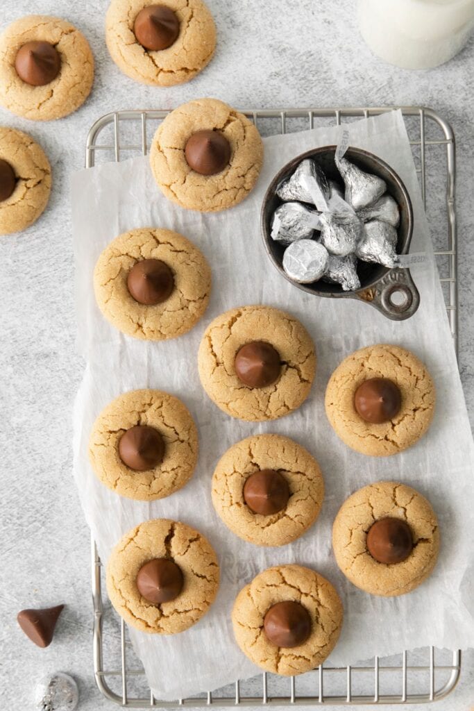 Peanut butter blossom cookies on a wire rack with parchment paper on the counter with a little bowl of wrapped hershey kisses to the side.