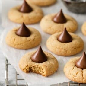 Peanut butter blossoms on a wire rack with parchment paper with one with a bite taken out of it so you can see the soft, chewy, texture of the cookie inside.