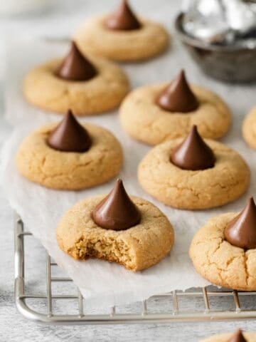Peanut butter blossoms on a wire rack with parchment paper with one with a bite taken out of it so you can see the soft, chewy, texture of the cookie inside.