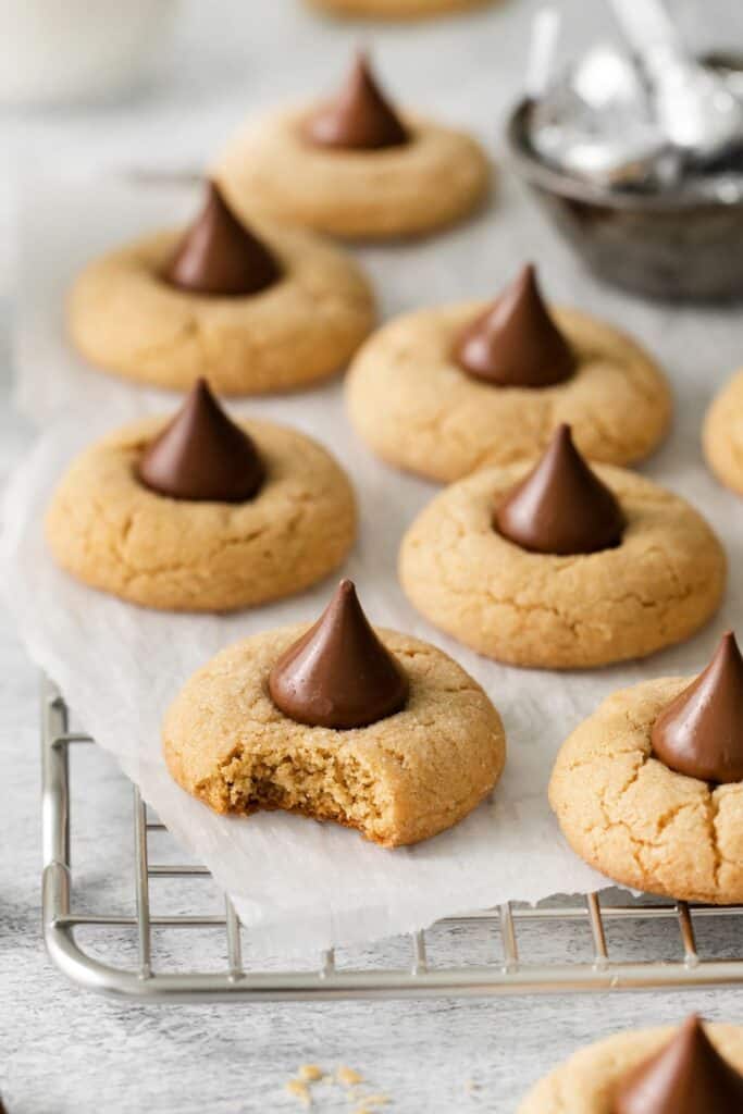 Peanut butter blossoms on a wire rack with parchment paper with one with a bite taken out of it so you can see the soft, chewy, texture of the cookie inside.