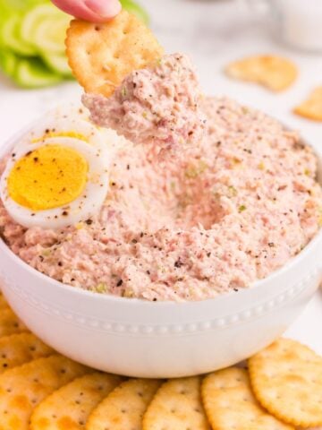 Ham salad in a bowl surrounded by crackers. A hand is scooping up ham salad with a cracker.