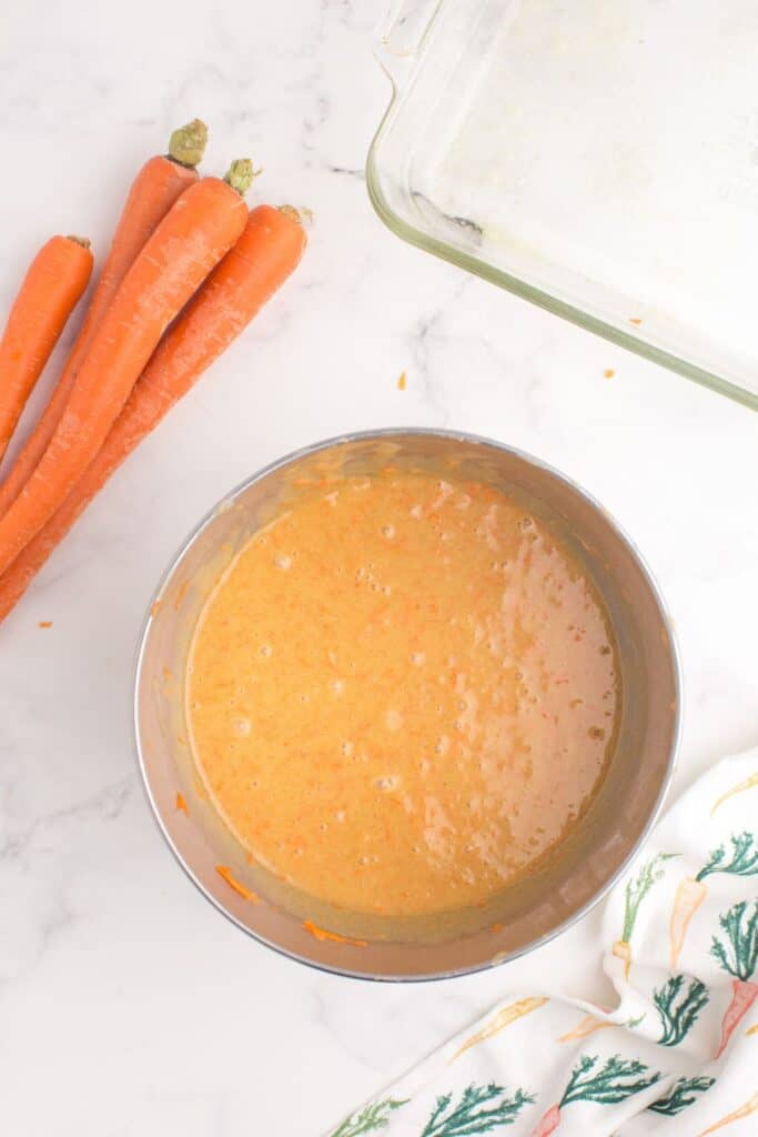 Carrot cake batter in a metal mixing bowl next to a prepped glass baking pan and a bunch of fresh carrots.
