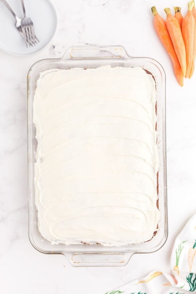 Frosted carrot cake in a glass baking pan. Cake is surrounded by white plates, forks, carrots, and a decorated tea towel.