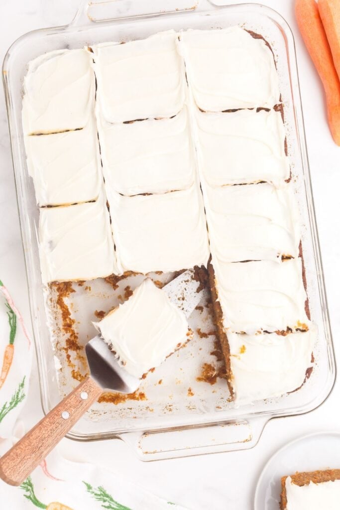 A glass baking dish with frosted carrot cake. A serving spatula has a slice of cake on it.