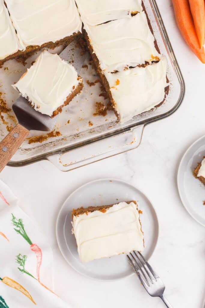 A glass 9x13" baking dish with sliced carrot cake in it. A white plate is next to the baking dish with a square slice of carrot cake on it.