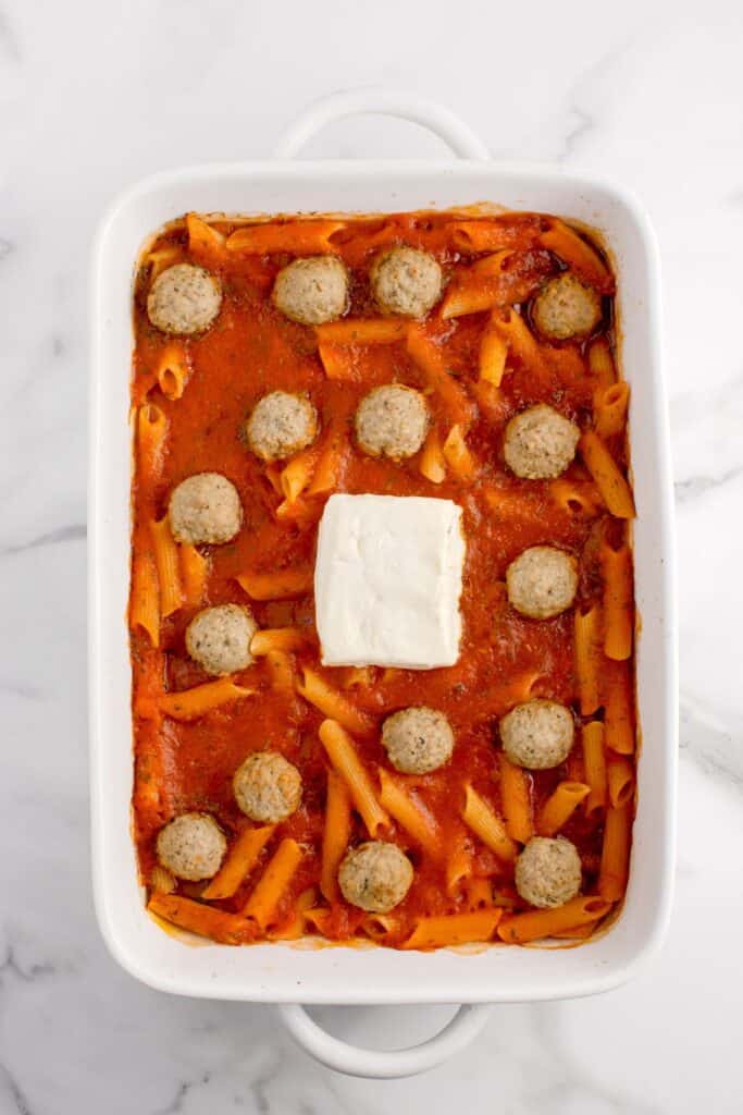 Process photo of pasta recipe. Baking dish is layered with penne pasta, red sauce, meatballs, and a partial block of cream cheese in the center.