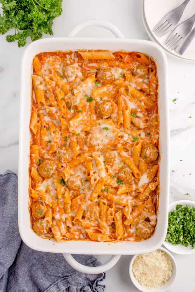 Baked penne pasta dish in a white baking dish. Casserole dish is surrounded by small bowls of parsley, parmesan cheese, a stack of plates with forks, fresh parsley, and a towel.