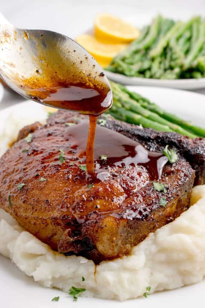 White dinner plate with a golden brown pork chop on a bed of mashed potatoes. A spoon is pouring the brown sugar sauce over the top.