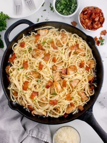 Cast iron pan of chicken carbonara. Pan is surrounded by small bowls of chopped bacon, parmesan, and parsley.