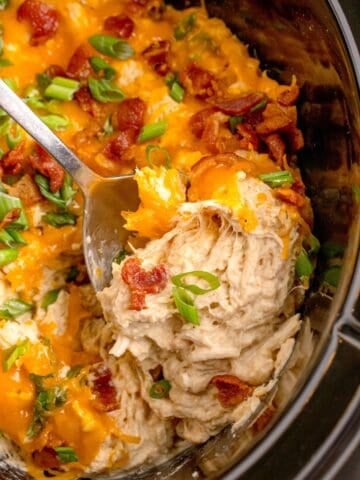 Crock pot insert with completed crack chicken. Chicken is shredded and layered with cheddar cheese, bacon, and green onions. A serving spoon is lifting a serving out of the insert.