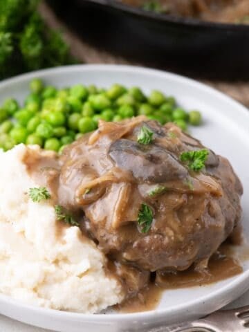 White dinner plate with hamburger steak. Served with mashed potatoes and peas.