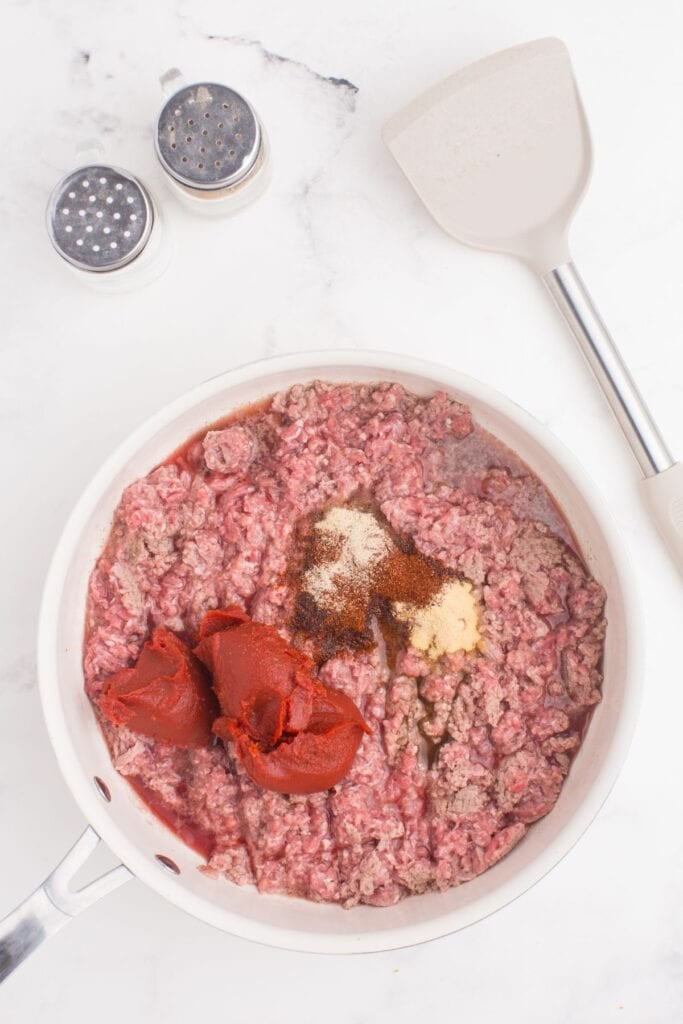 Skillet with partially cooked ground beef, seasonings, and tomato paste. Salt and pepper shakers sit above the skillet along with a plastic spatula.
