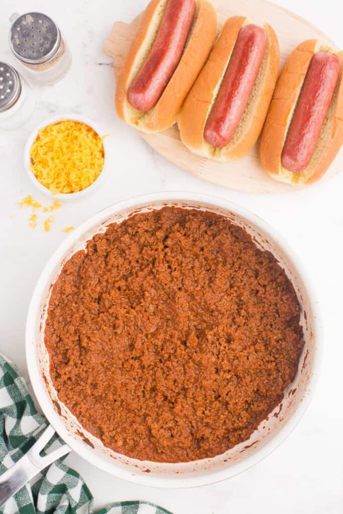 A platter of prepped hot dogs with a small bowl of shredded cheddar cheese and a skillet with homemade chili in it.