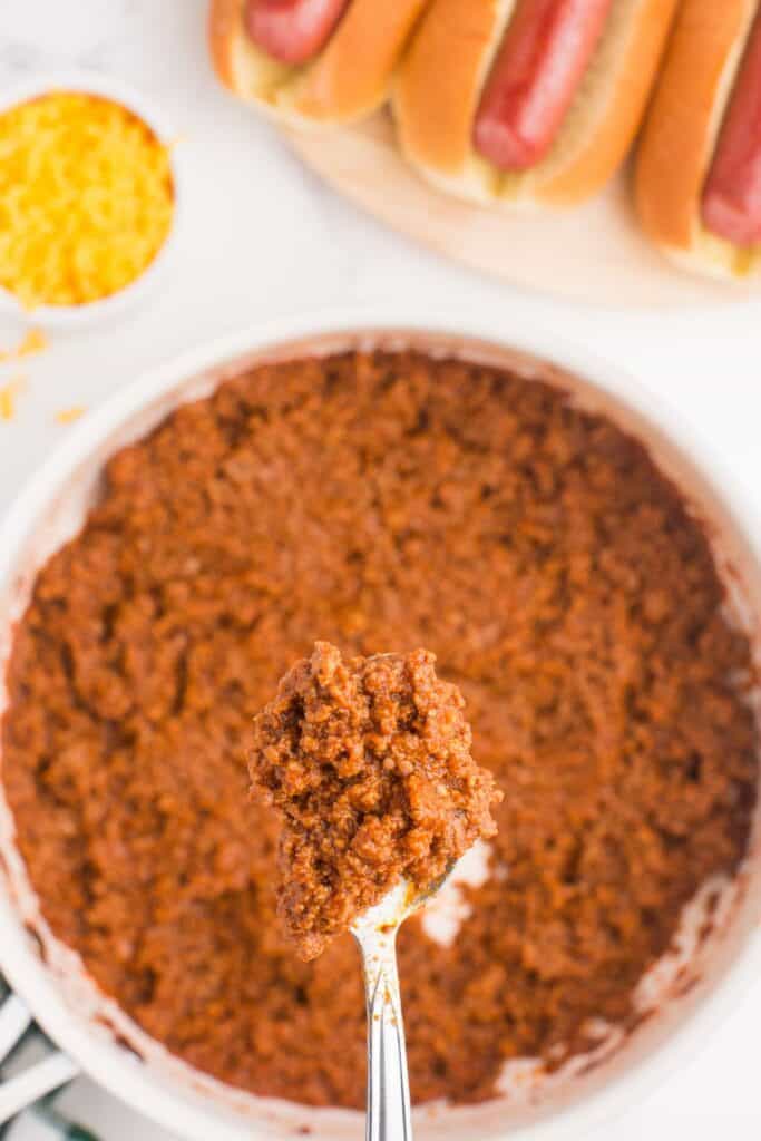 A silver spoon is lifting up a bite of homemade chili above a skillet. A small bowl of shredded cheese is next to the skillet, along with a platter of prepped hot dogs.