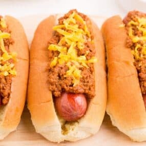 Close up view of a row of hot dogs on a serving platter with homemade chili and shredded cheddar cheese on top.