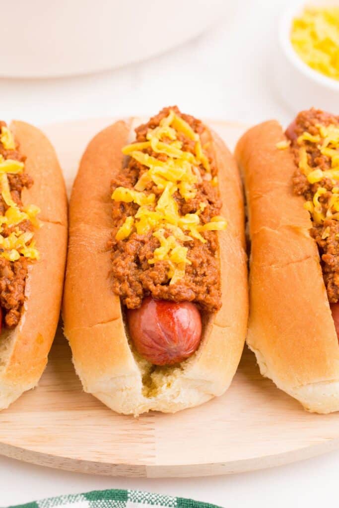 Close up view of a row of hot dogs on a serving platter with homemade chili and shredded cheddar cheese on top.