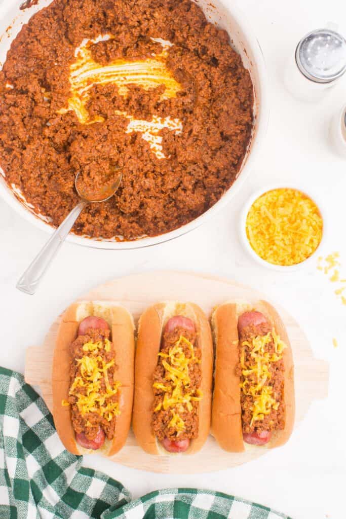 Row of hot dogs on a serving platter with homemade chili and shredded cheddar cheese on top. A skillet with homemade chili sits above the hot dogs along with a small bowl of shredded cheddar cheese.