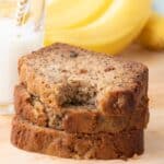 3 slices of banana bread in a stack. The top piece has a bite taken out of it. A glass of milk and bunch of bananas can be seen in the background.