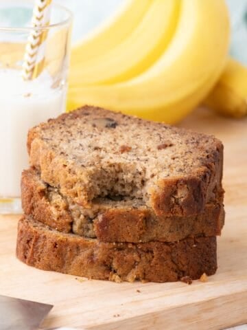 3 slices of banana bread in a stack. The top piece has a bite taken out of it. A glass of milk and bunch of bananas can be seen in the background.