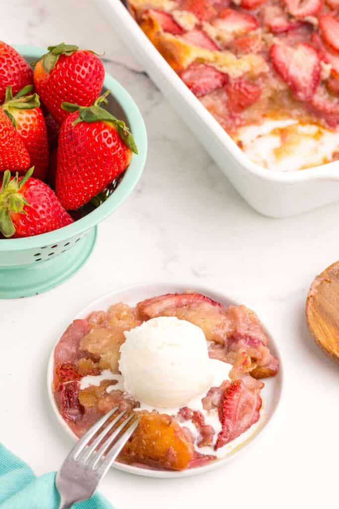 Small bowl of cobbler with a scoop of ice cream on top. Bowl of strawberries and baking dish with strawberry cobbler behind the bowl.
