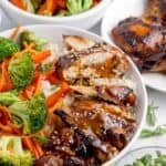 Teriyaki bowl surrounded by a bowl of veggies, a small glass bowl of teriyaki sauce, a plate of chicken thighs, and a small bowl of chopped green onions.