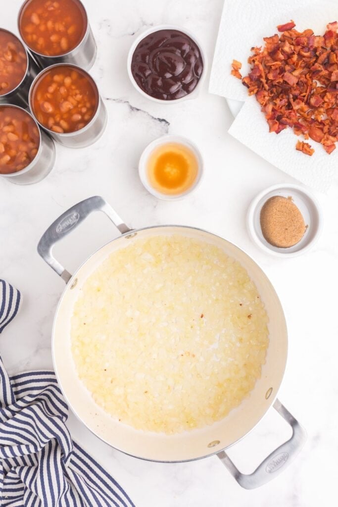 A pot with chopped onion at the bottom next to 4 cans of baked beans, a small bowl of apple cider vinegar, a bowl of BBQ sauce, a plate of chopped bacon, and a small bowl of packed dark brown sugar.