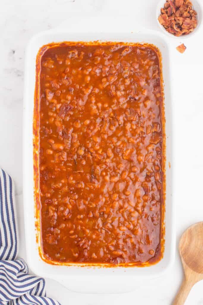 A white baking dish with baked beans in it, next to a wooden serving spoon, and a small bowl of chopped bacon.