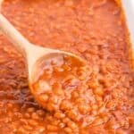 White baking dish with baked beans in it. A wooden serving spoon is lifting up a serving of beans.