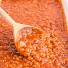 White baking dish with baked beans in it. A wooden serving spoon is lifting up a serving of beans.