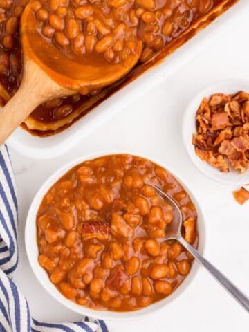 A white bowl of baked beans with a metal spoon. Beside the bowl is a small bowl of chopped bacon and the baking dish of baked beans with a wooden serving spoon in it.