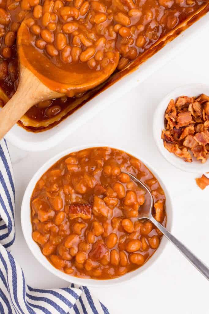 A white bowl of baked beans with a metal spoon. Beside the bowl is a small bowl of chopped bacon and the baking dish of baked beans with a wooden serving spoon in it.