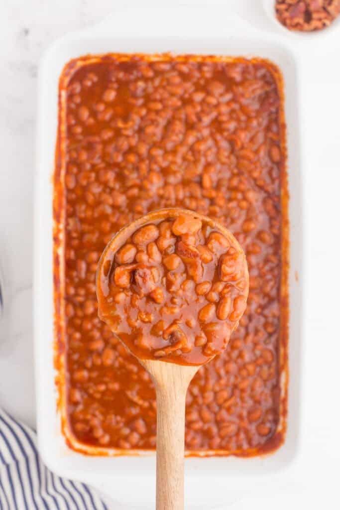 A wooden spoon lifting up a serving of baked beans above a baking dish.