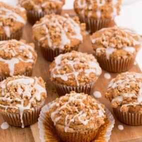 Rows of coffee cake muffins lined up on a wooden cutting board. The muffin in front have the paper wrapping peeled down.