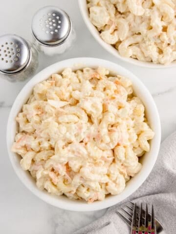 Two white bowls of macaroni salad. Bowls are flanked by a tea towel, forks, salt and pepper shakers.