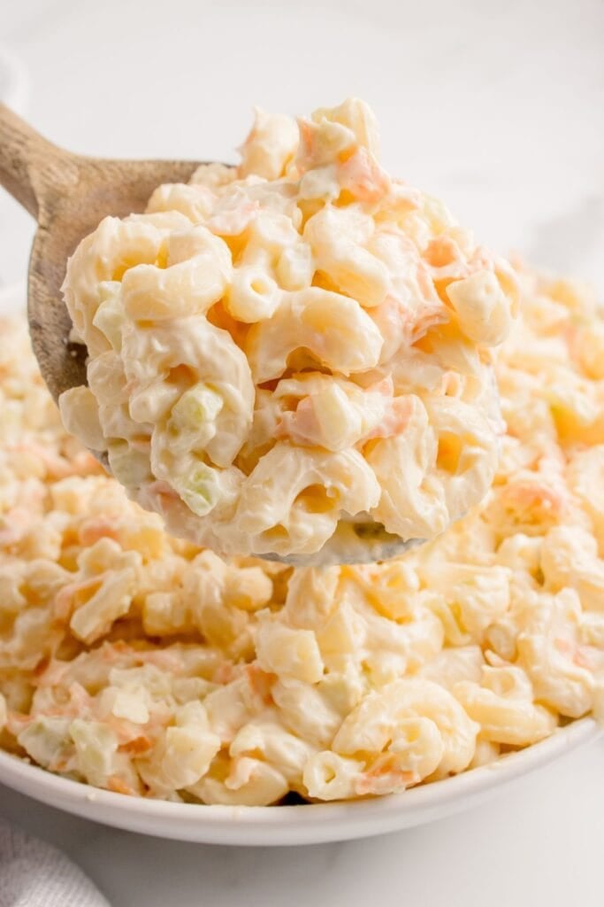 Creamy macaroni salad in a serving bowl with a wooden serving spoon lifting a portion above the bowl.