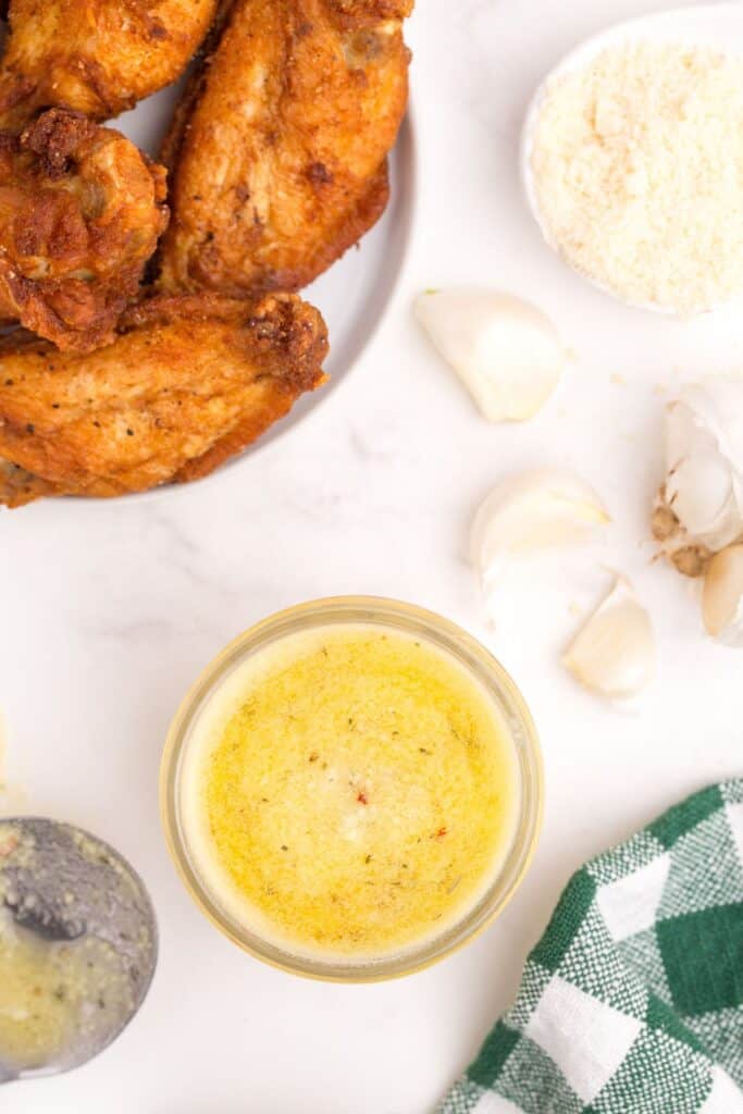 Small bowl of sauce next to a spoon, a plate of chicken wings, a small bowl of grated parmesan, and several cloves of garlic.