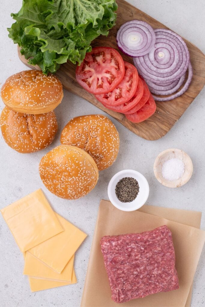 Ingredients for hamburgers in the oven: ground beef, salt, black pepper, American cheese, hamburger buns, lettuce leaves sliced tomatoes, and onions.
