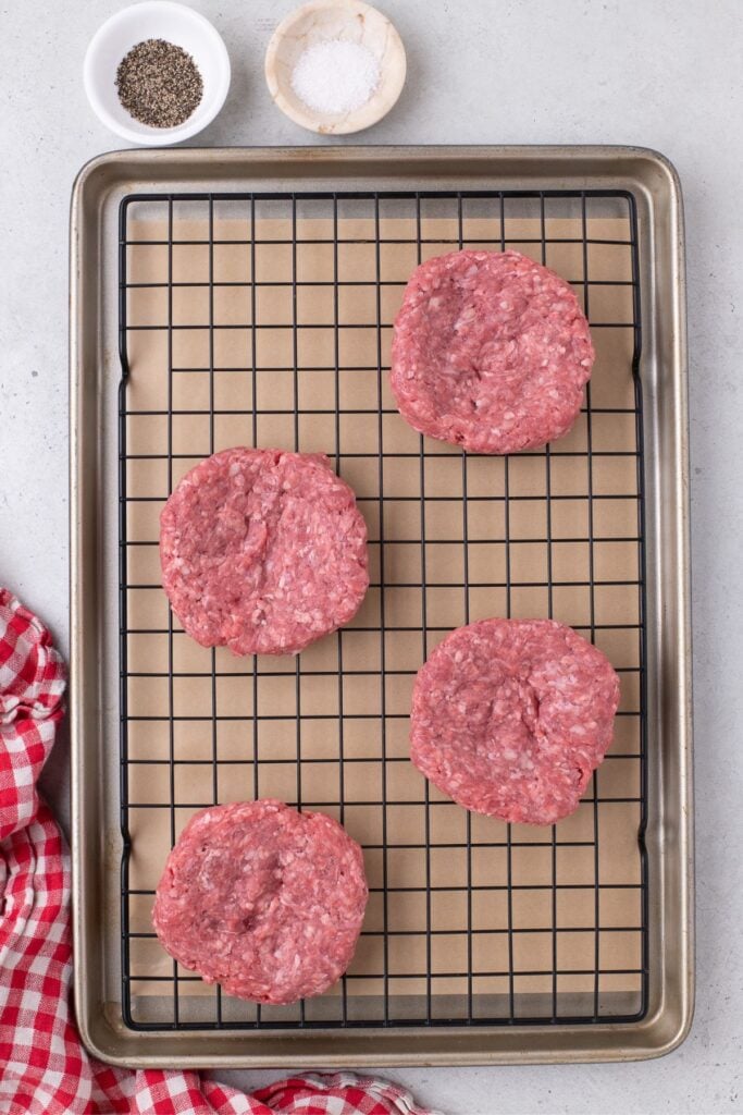 Raw ground beef hamburger patties are lined on a baking sheet. Small bowls of salt and pepper sit just above the baking sheet.