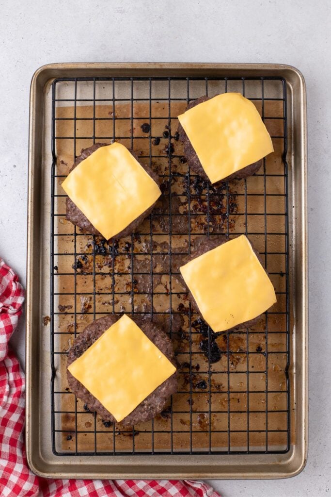 View of four cooked ground beef patties from above. Patties all have melted cheese, and are sitting on a wire rack on the baking sheet. The baking sheet is lined with parchment.