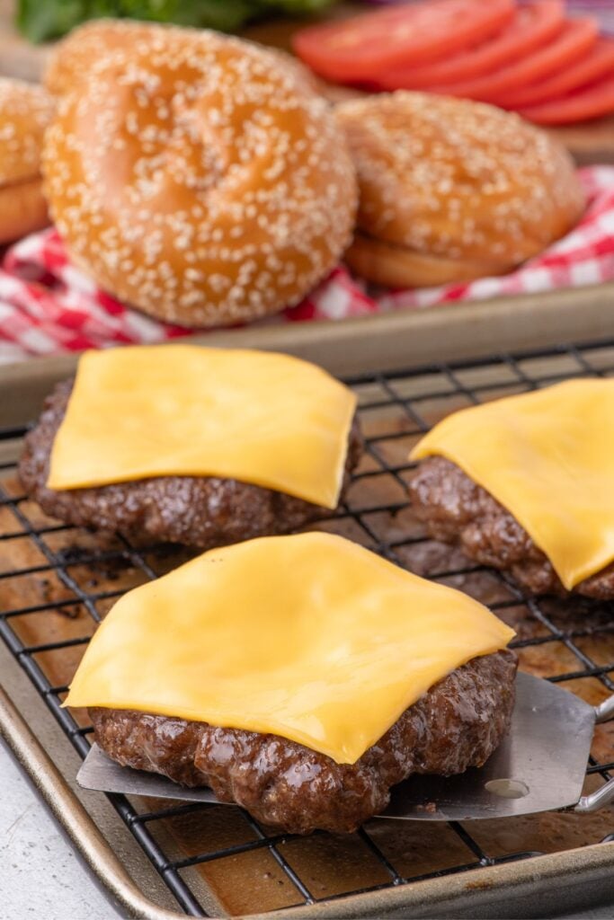 Cooked hamburger patties with melted cheese on a baking sheet. Hamburger buns and sliced vegetables are in the background.