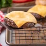Metal spatula lifting a beef burger patty with melted cheese off a baking sheet with a wire rack.