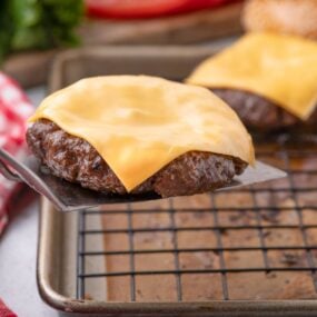 Metal spatula lifting a beef burger patty with melted cheese off a baking sheet with a wire rack.