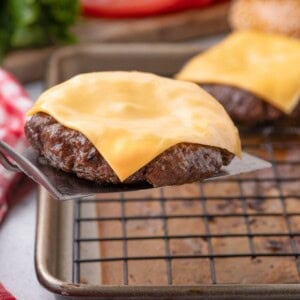 Metal spatula lifting a beef burger patty with melted cheese off a baking sheet with a wire rack.