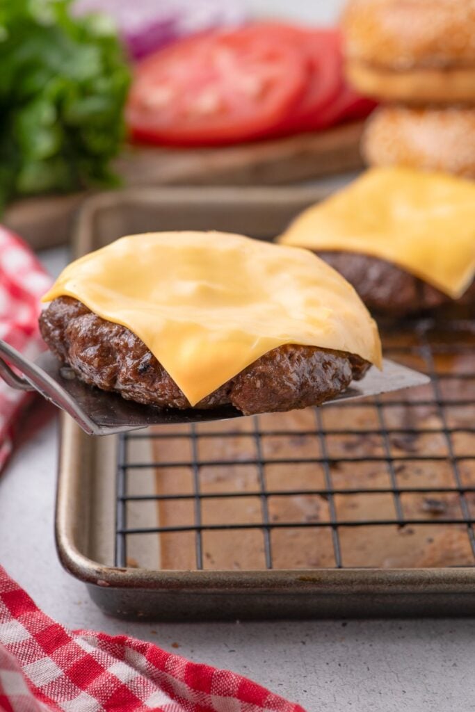Metal spatula lifting a beef burger patty with melted cheese off a baking sheet with a wire rack.