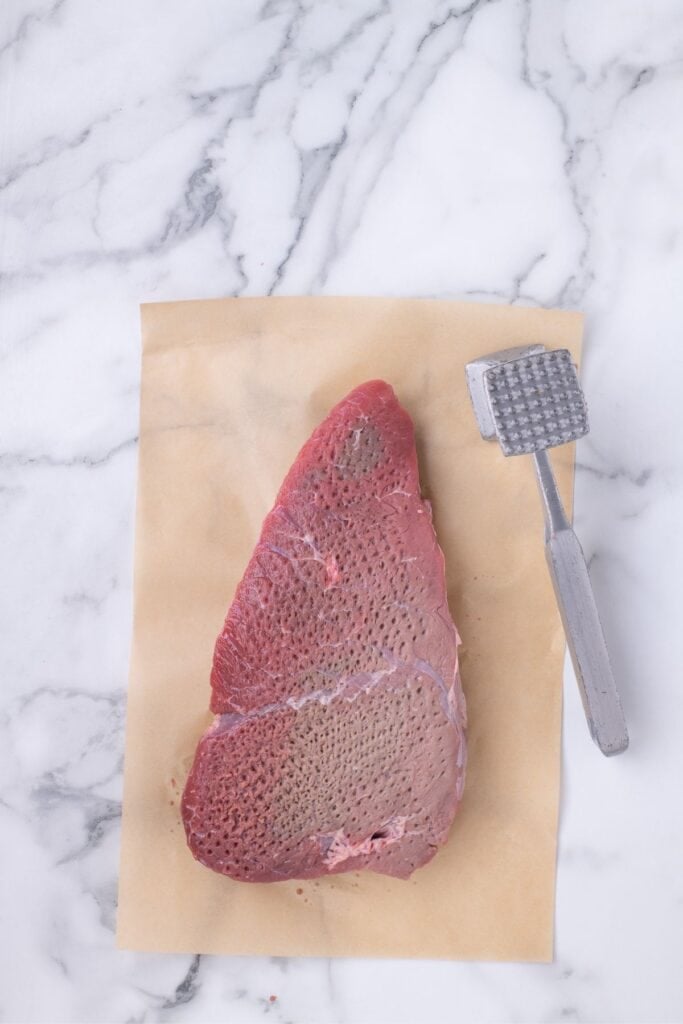 Raw London broil roast on a sheet of parchment paper. It has been pierced with a meat tenderizer mallet several times across the front.