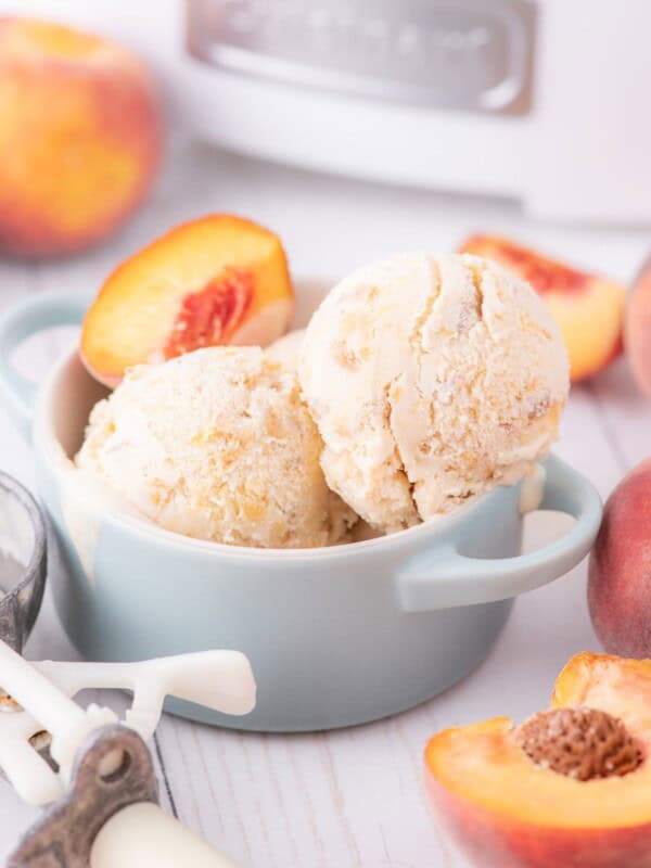 Ice cream scooped into a bowl garnished with a fresh peach slice. The bowl is surrounded by fresh whole peaches, peach slices, and an ice cream scoop.