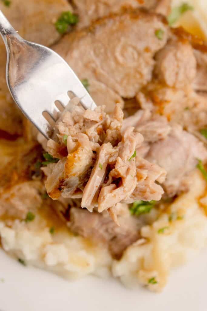 Forkful of pork roast is lifted above a white dinner plate with pork roast and mashed potatoes on it.