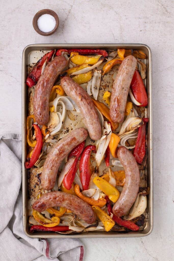 Sheet pan with cooked Italian sausage, bell peppers, and onions. A small pinch bowl with salt is positioned above the sheet pan.
