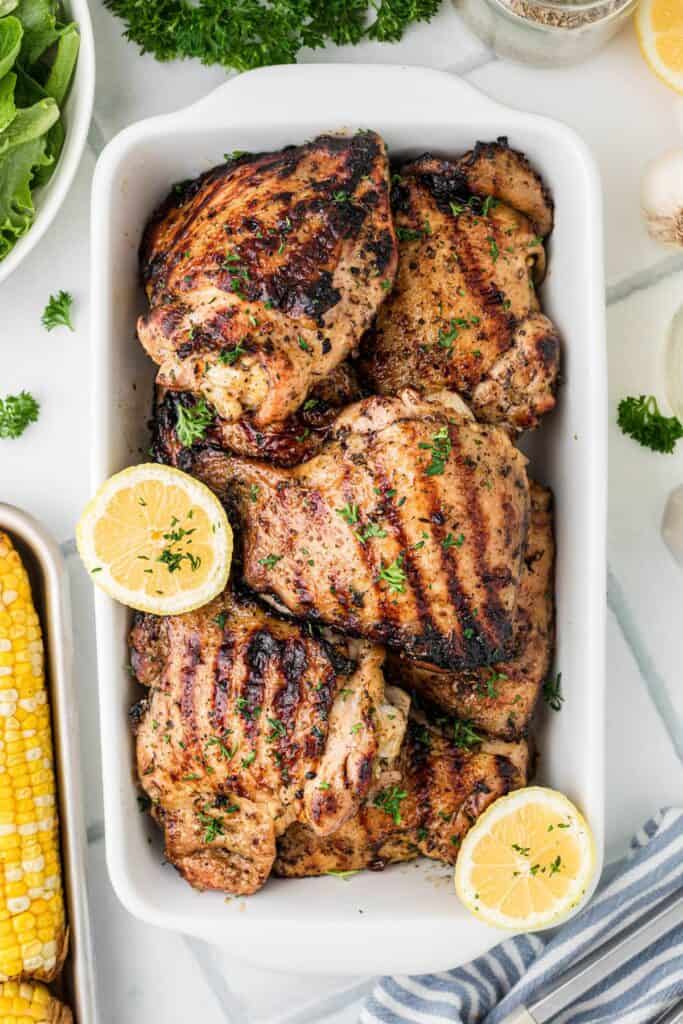 A white baking dish with several grilled chicken thighs piled into it. A sliced lemon is balanced on the sides of the dish.