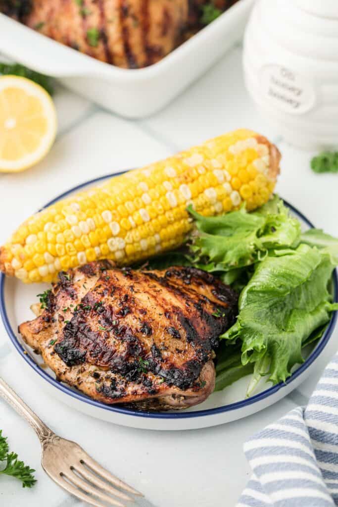 A white dinner plate with a blue rim filled with grilled chicken thighs, corn on the cob, and salad greens.